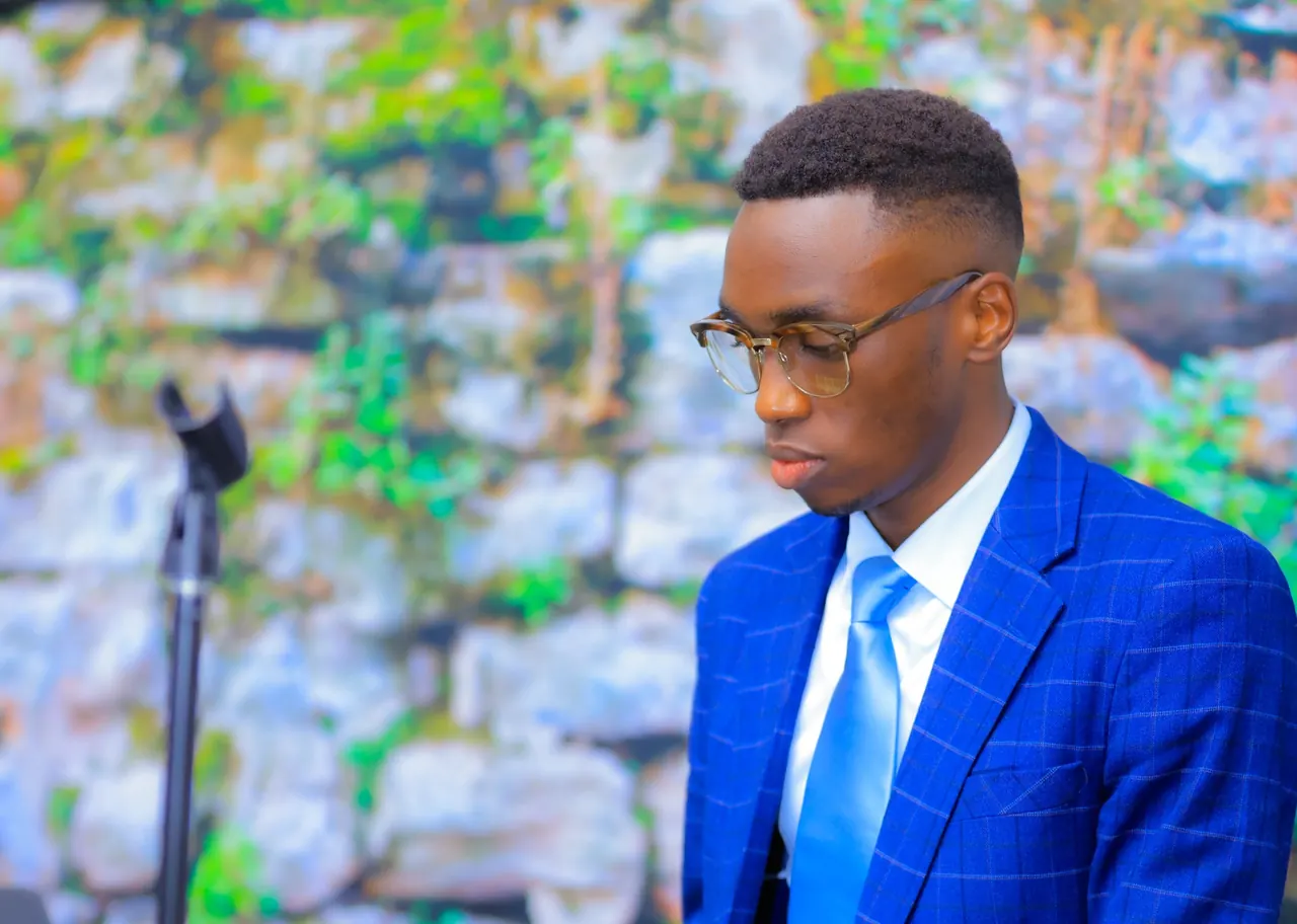 Fiston in a blue suit and glasses, outdoors in front of a stone wall.